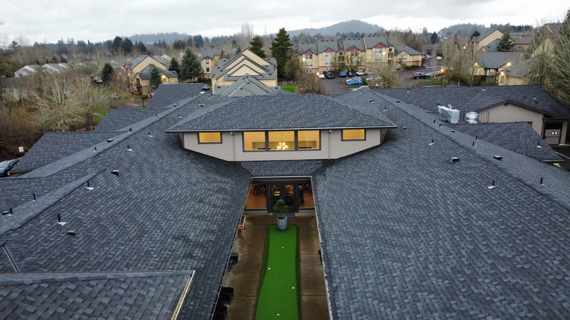 Overhead view of a building with dark gray roofs, a green putting green, and a residential neighborhood in the background.