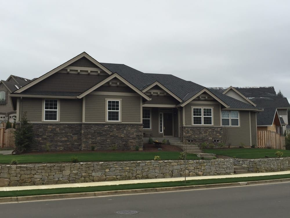 Tan and brown Craftsman-style house with stone accents, lawn, and retaining wall on a cloudy day.