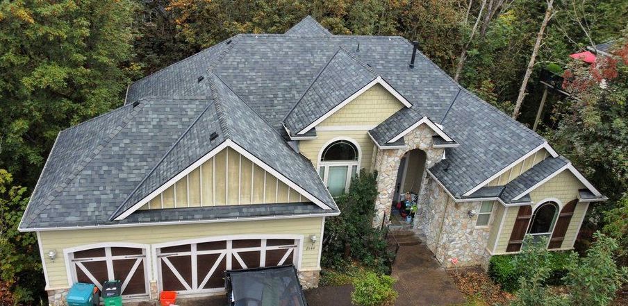 House with gray roof and tan siding surrounded by trees.