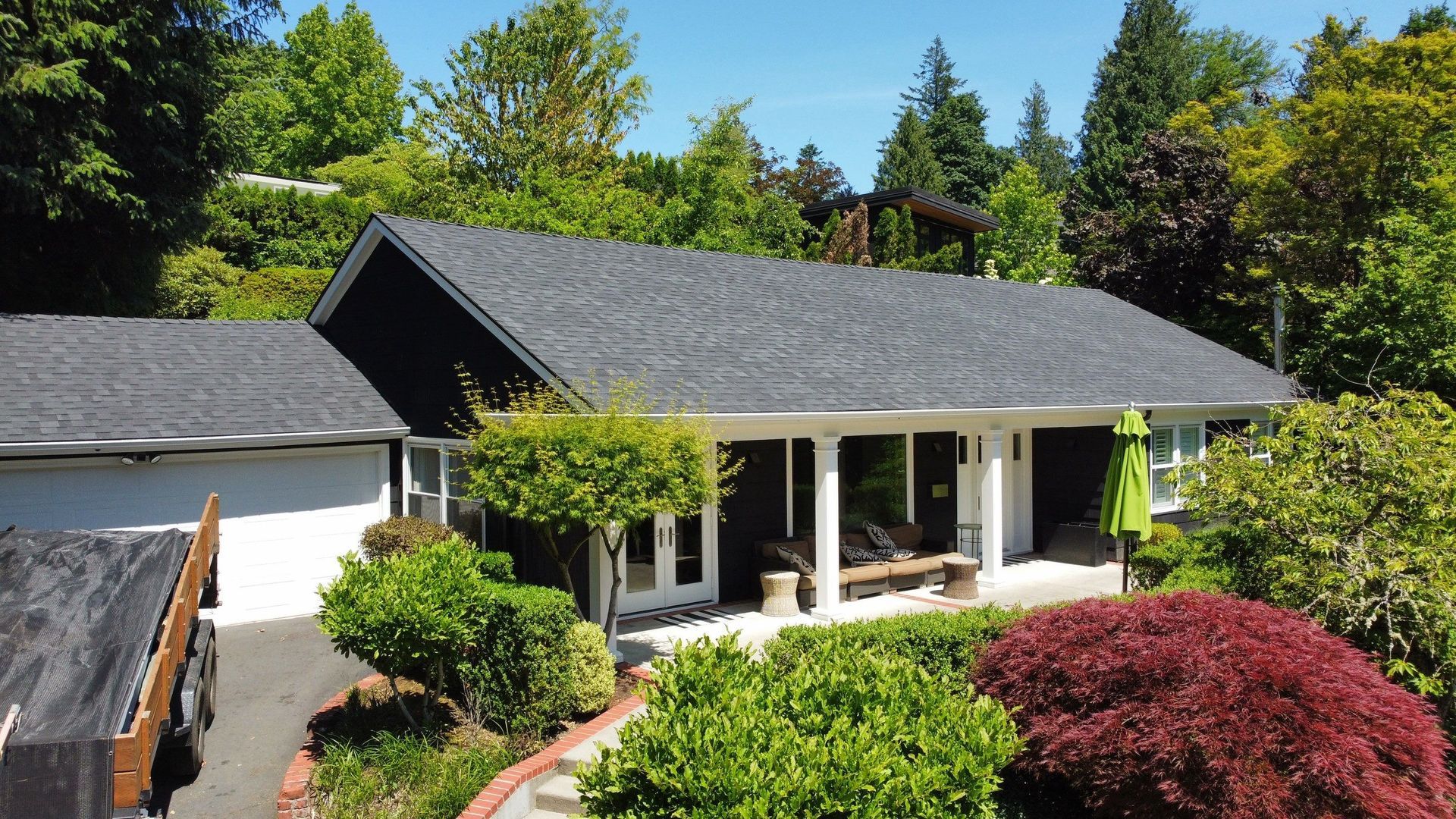 Residential house with dark gray roof, surrounded by green trees and bushes. Sunny day.