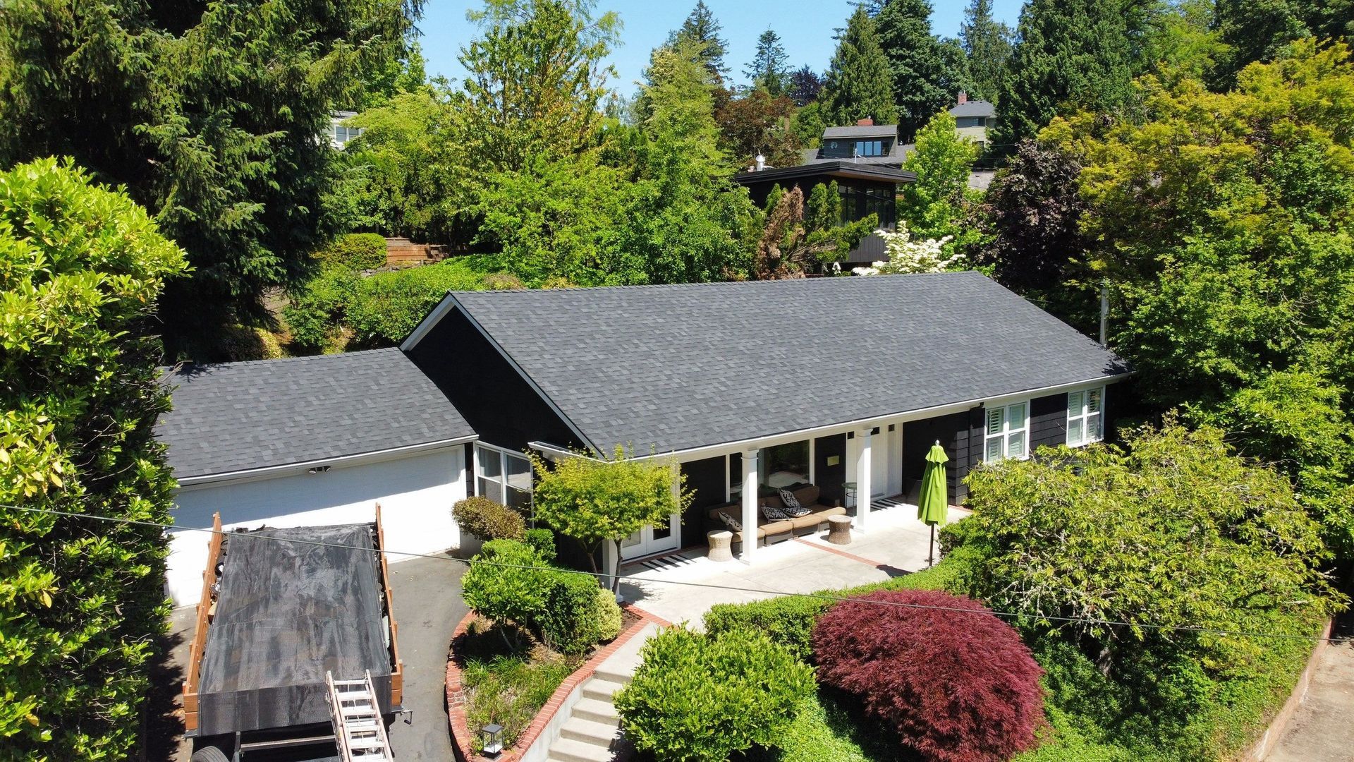 House with dark roof surrounded by lush green trees, a driveway, and a trailer parked next to the garage.