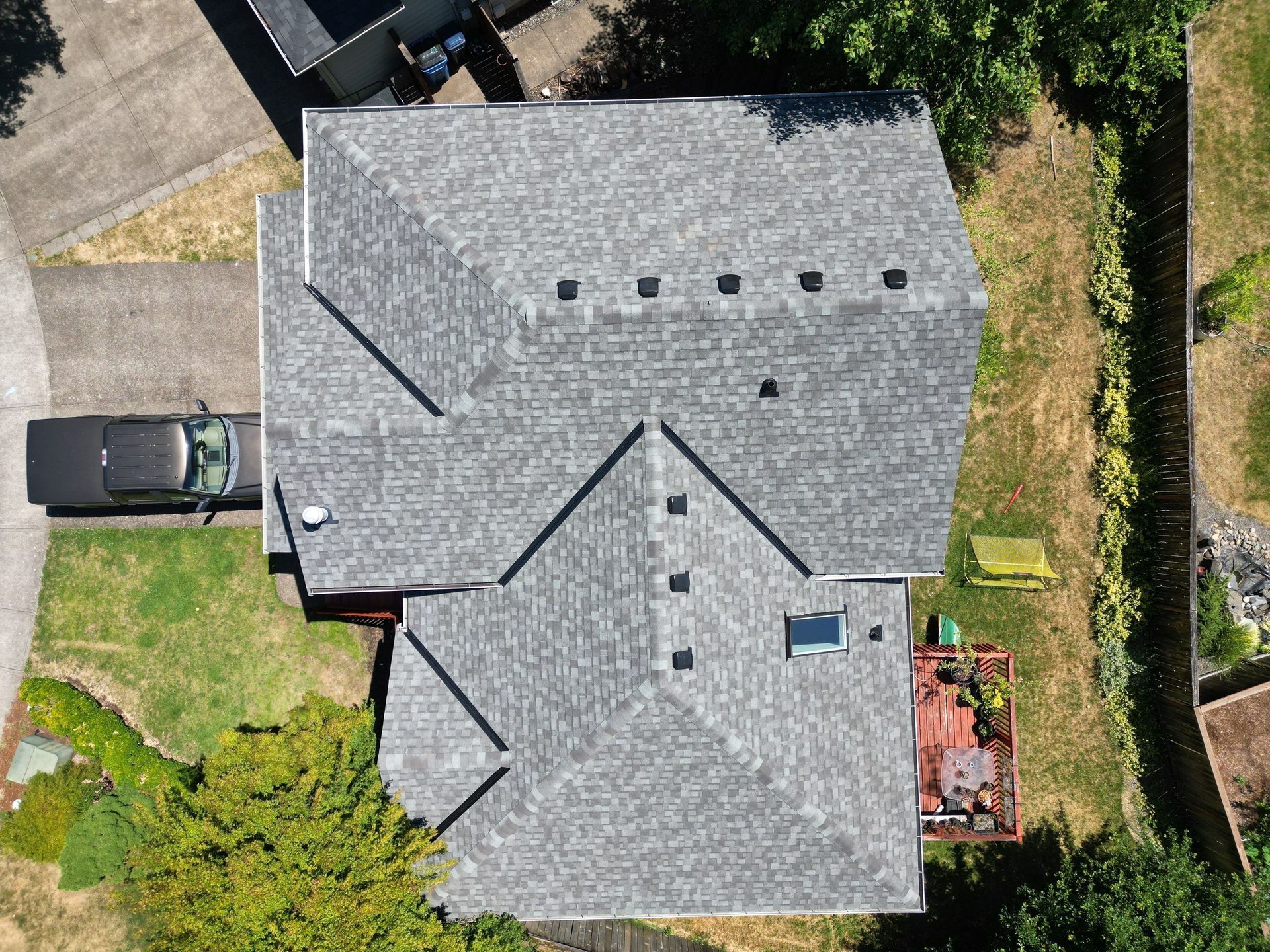 Overhead view of a house with a gray shingle roof, black car in driveway, surrounded by green grass and trees.