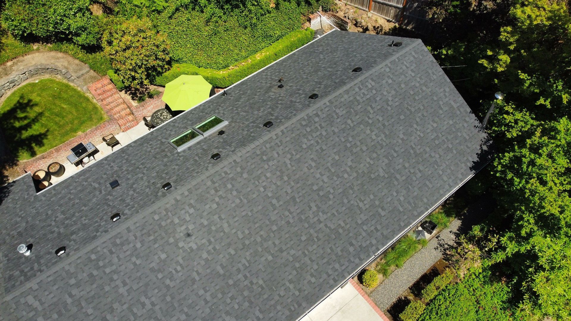 Aerial view of a dark gray shingled roof with vents, surrounded by trees and a small patio.