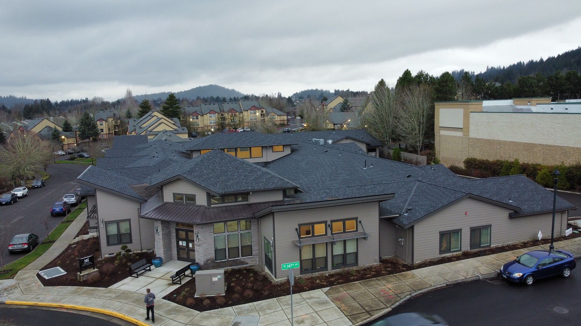 Exterior view of a light gray building with a dark roof on a cloudy day, with a person and cars nearby.
