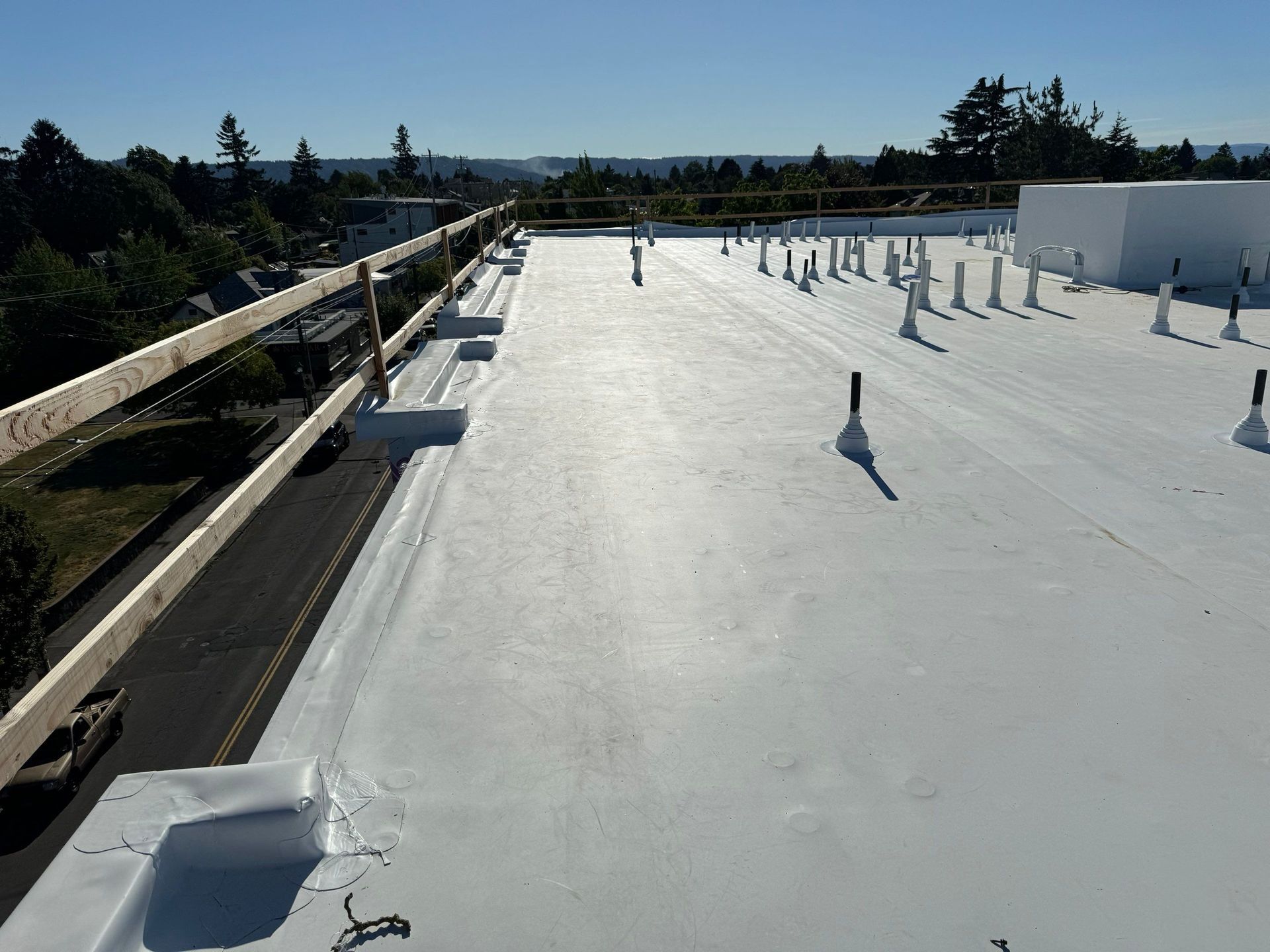 White flat roof with safety railing, pipes, and view of houses and trees.
