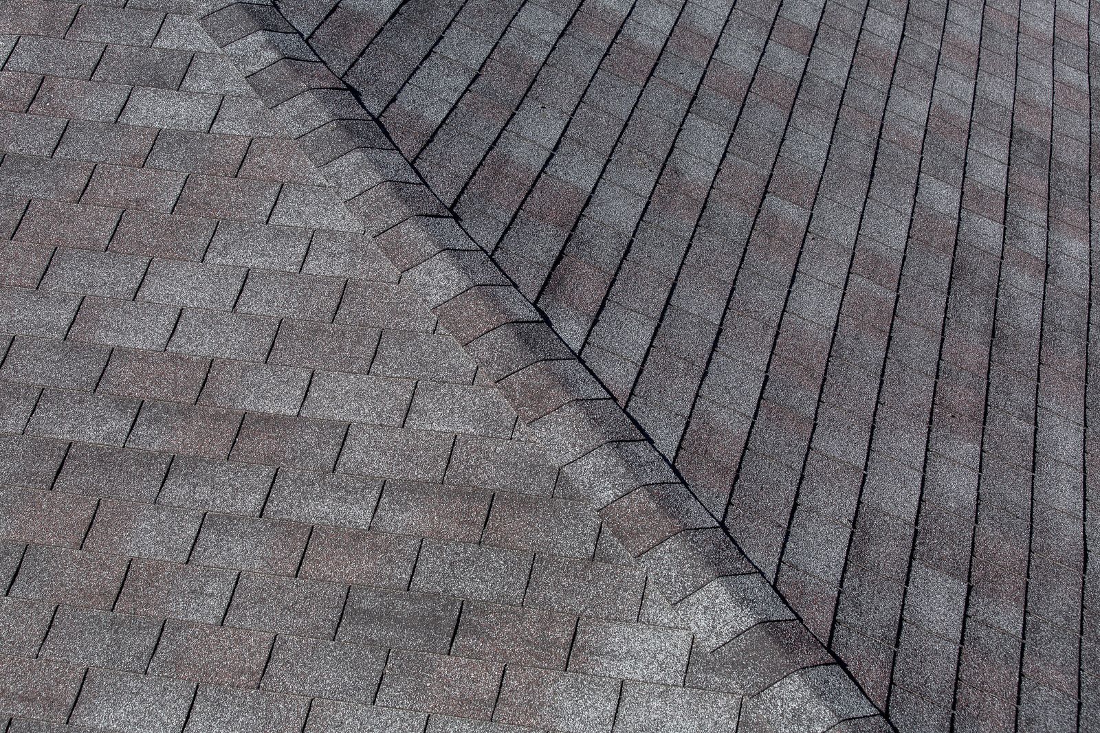 Close-up view of a roof with gray and brown asphalt shingles; a roof intersection is visible.