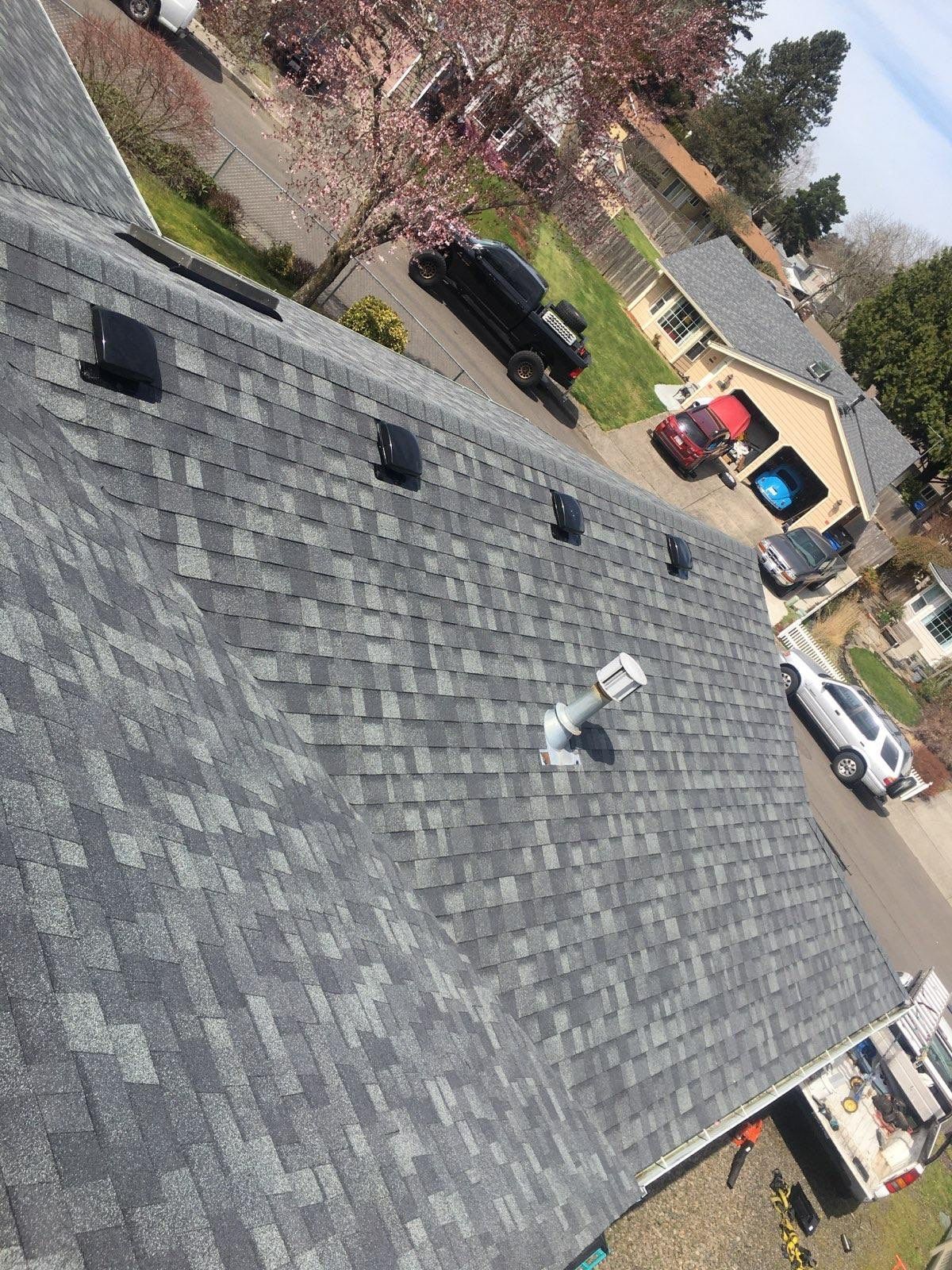 Gray shingle roof with black vents, overlooking a neighborhood with houses and cars.