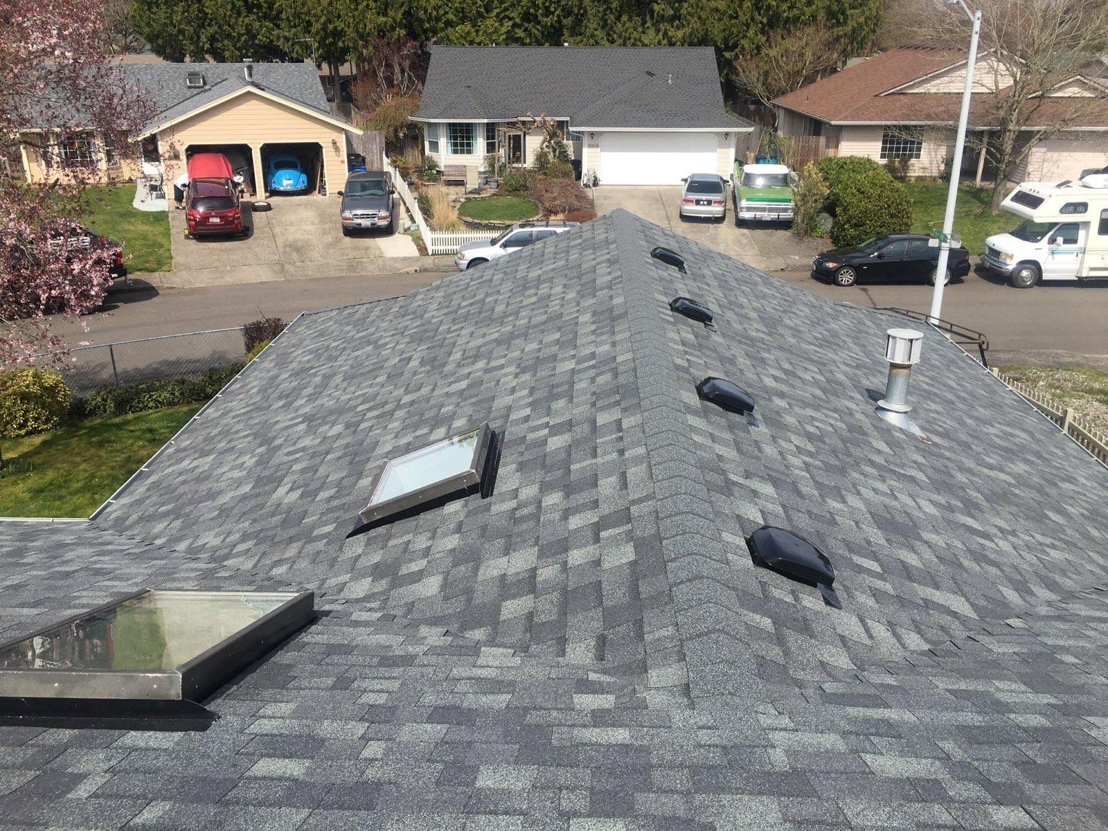 A house roof with gray shingles, skylights, and vents. Other houses and parked cars in the background.
