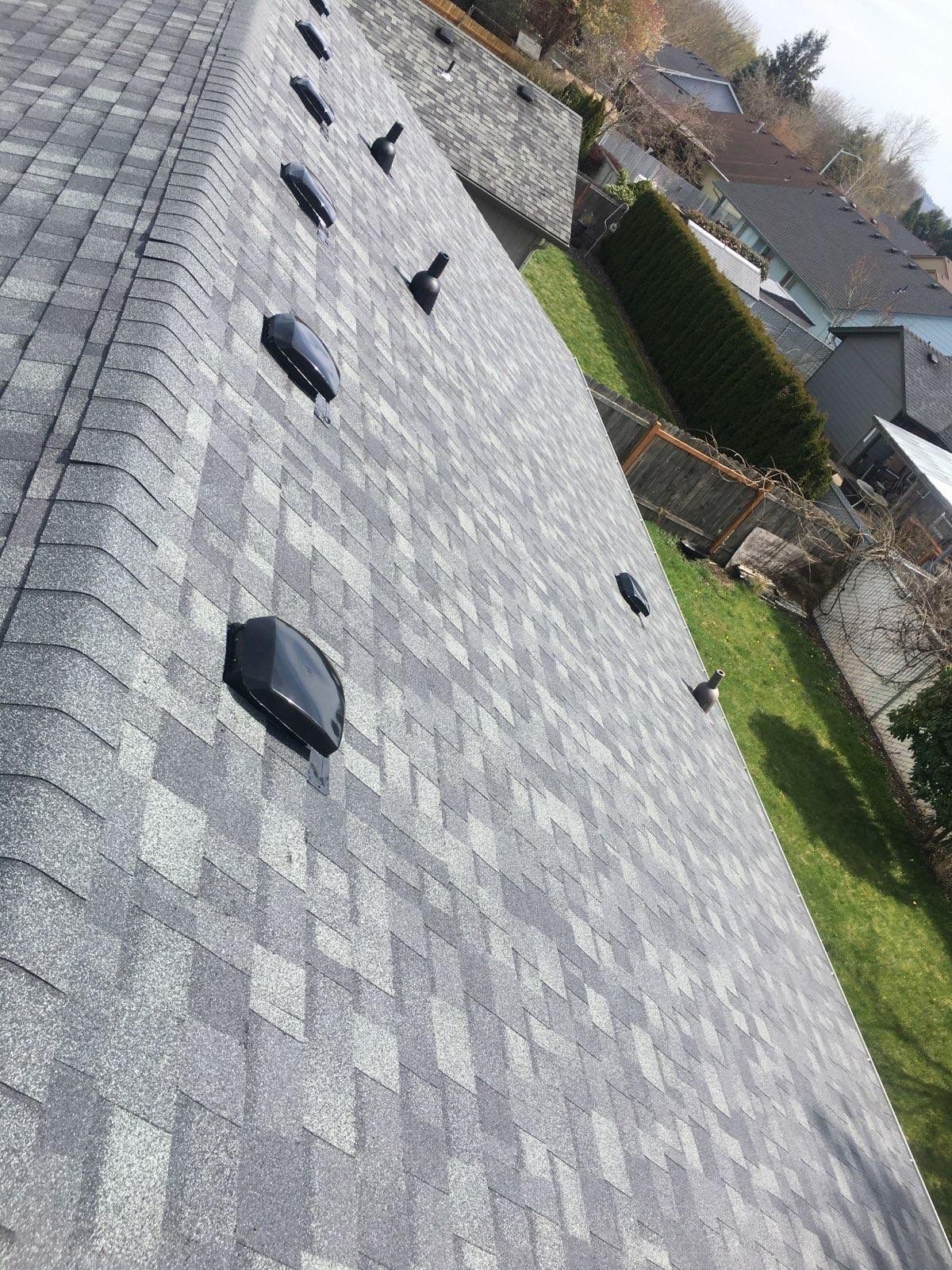 Gray shingled roof with several black vents, sloping downwards over a lawn and other houses.