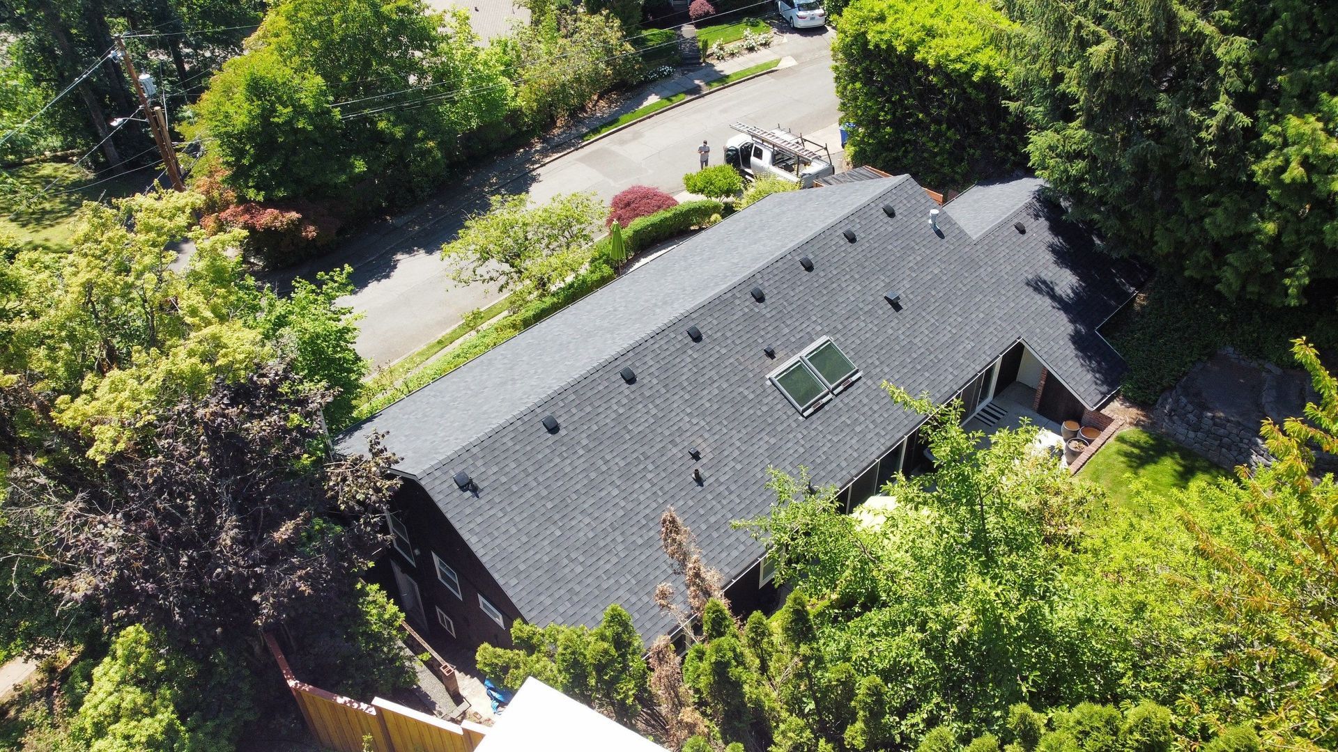 Aerial view of a dark-roofed house surrounded by trees, with a driveway in a suburban setting.