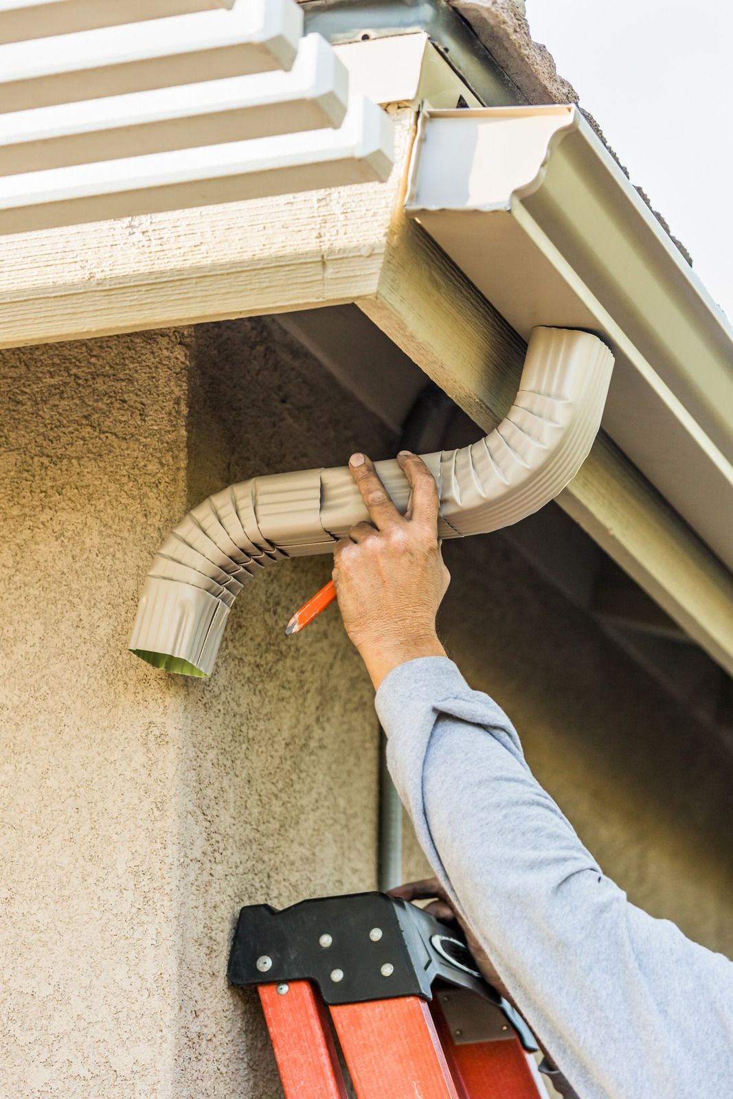 Person on ladder installing a tan gutter downspout on a beige stucco house.
