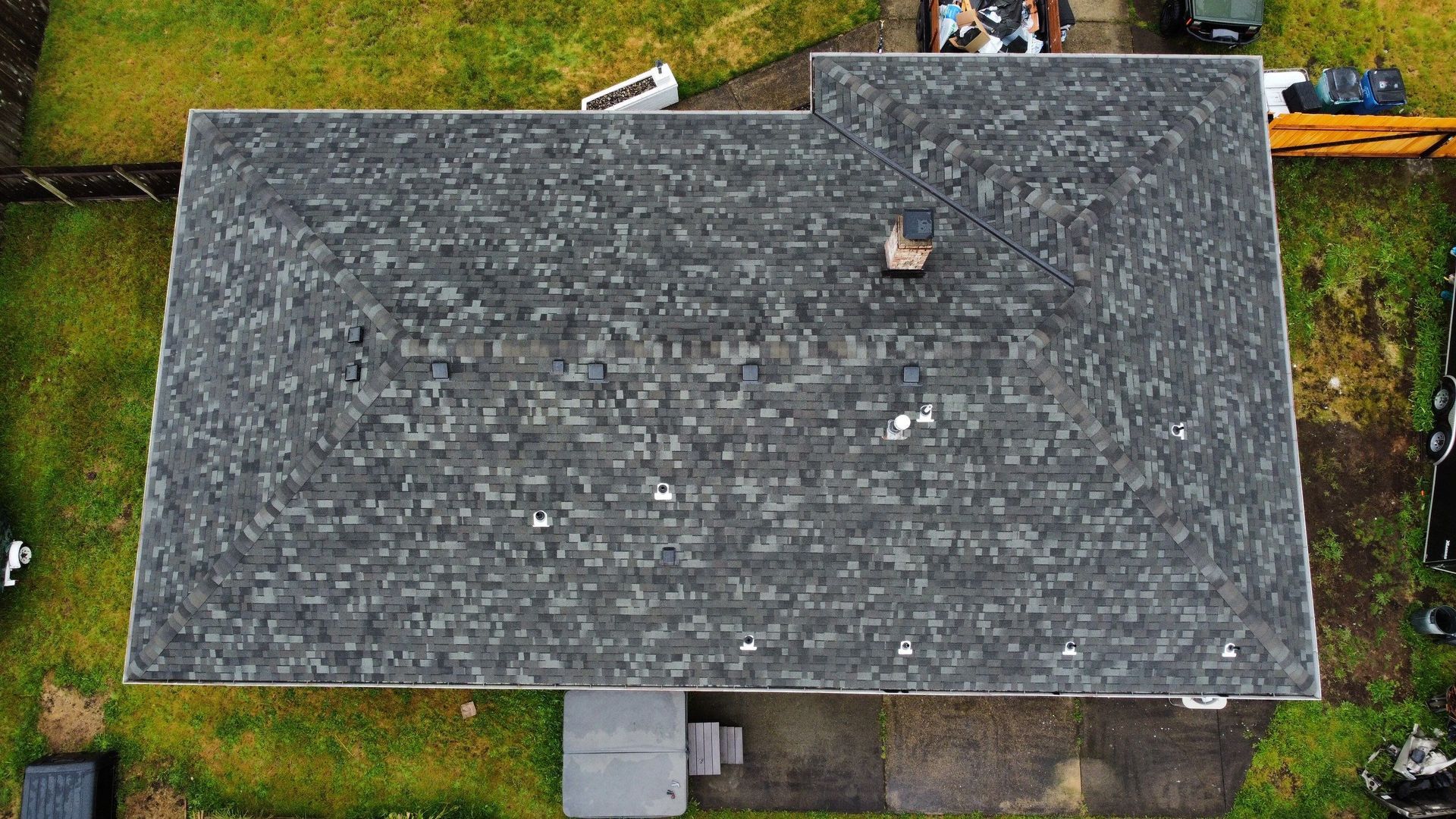 Overhead view of a gray shingled roof with a chimney and surrounding yard.