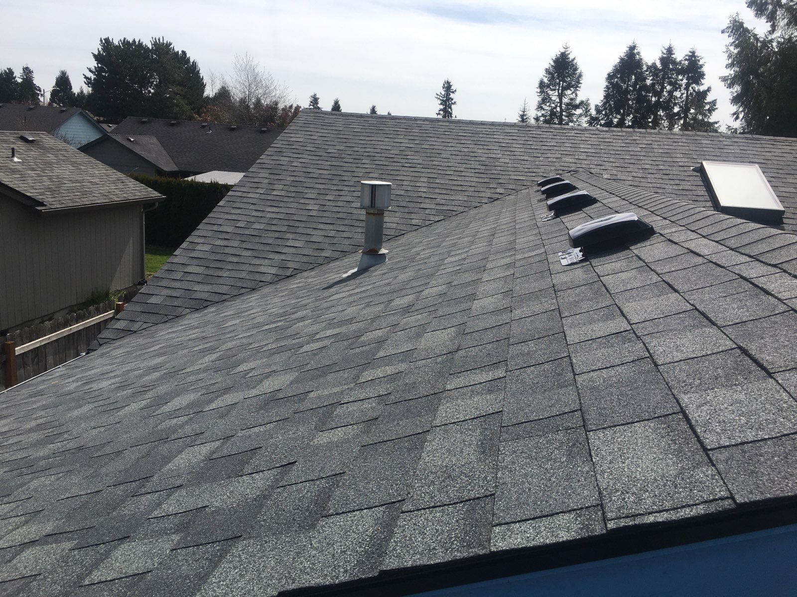 Gray asphalt shingle roof with vents and a chimney on a sunny day in a suburban neighborhood.