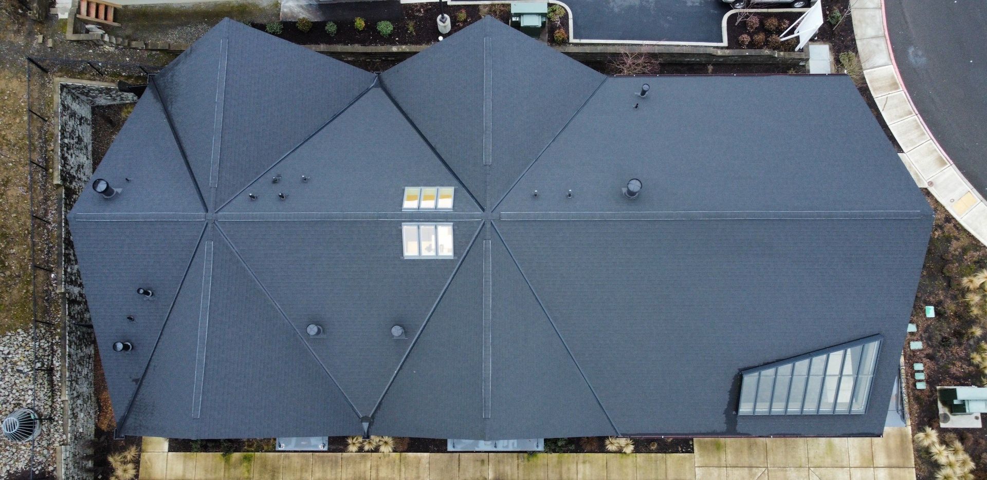 Overhead view of a modern house with a complex dark gray geometric roof, small windows, and a long driveway.