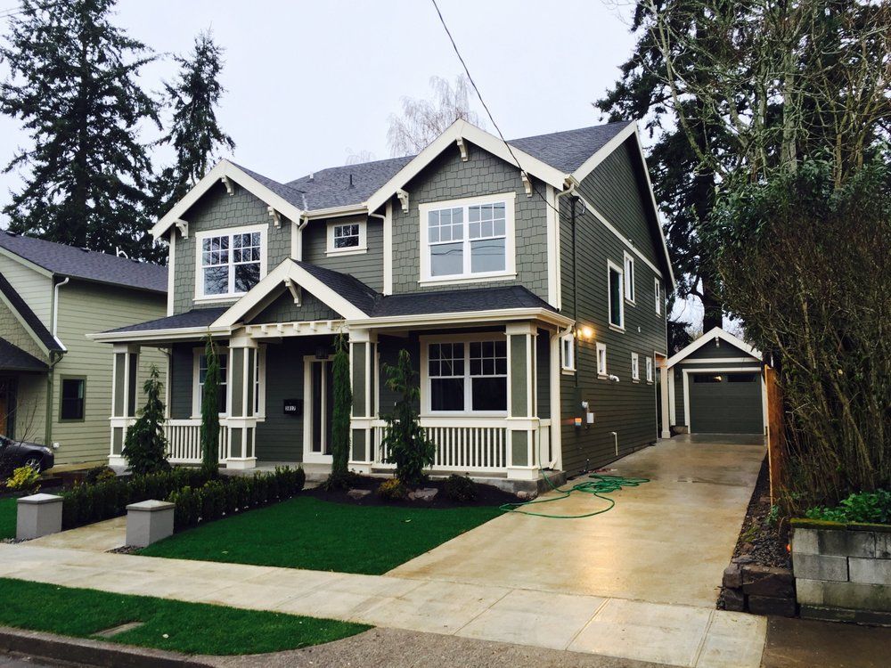 Green two-story house with white trim and a gray roof, flanked by grass and a driveway.