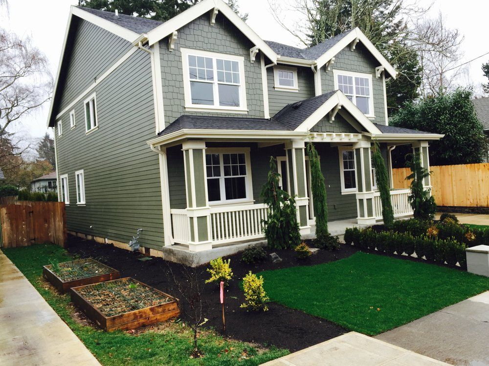 Green two-story house with white trim and front porch, neat lawn, and small garden beds.