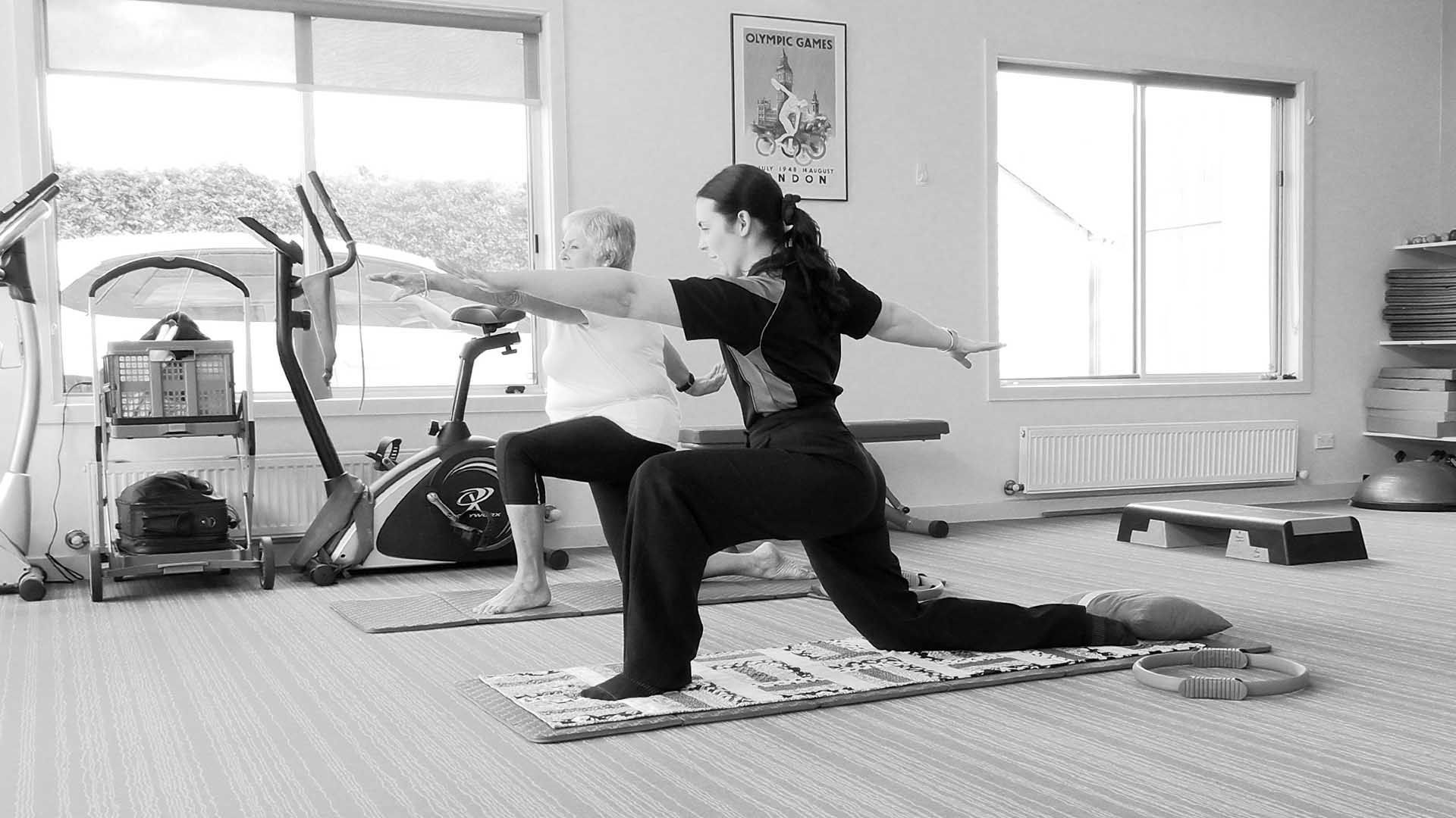 Two people doing yoga poses in a bright room with exercise equipment.