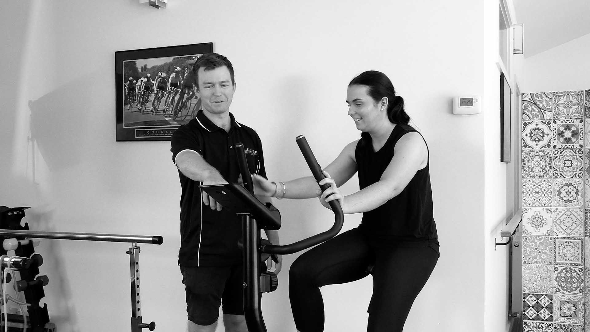 Man assisting woman on exercise bike, indoors. Both smiling.