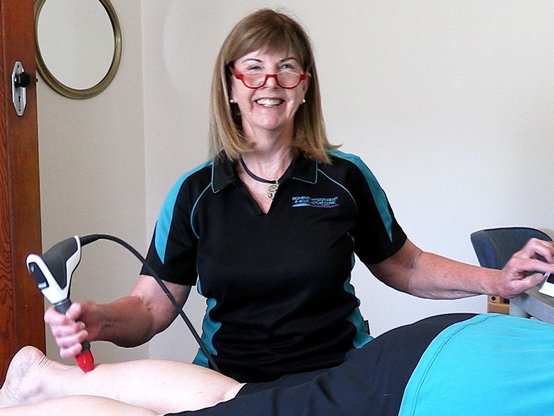 Woman with glasses using a therapy device on a patient's leg. She is smiling in an office setting.