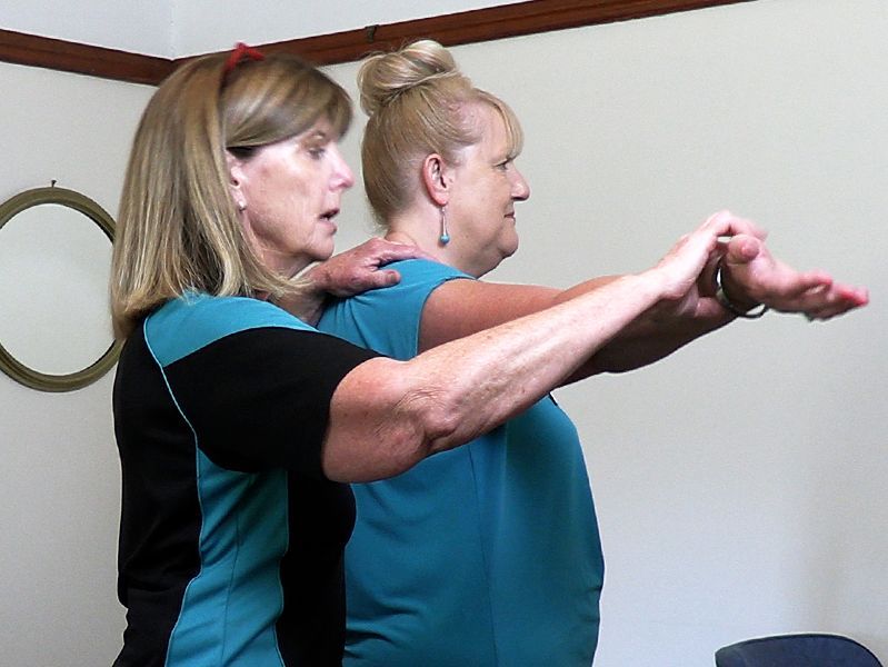 Woman assists another, guiding her arms forward, both in a room.