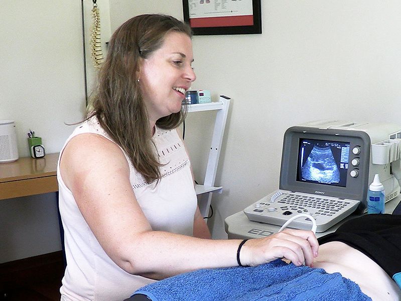 Woman using ultrasound machine on a patient, smiling in a medical setting.