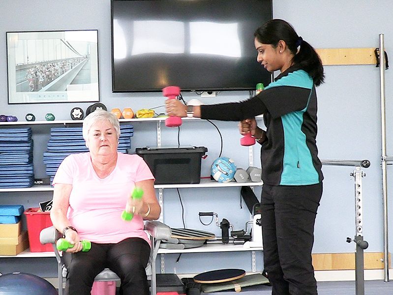 Woman in pink shirt exercises with weights, guided by a trainer in a therapy room.