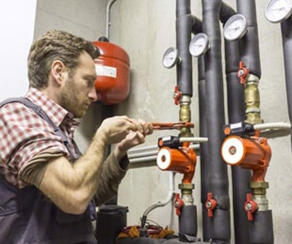 A Person in Work Clothes Adjusting Valves on Heating Pipes in A Utility Room — Weav Plumbed in Emerald, QLD