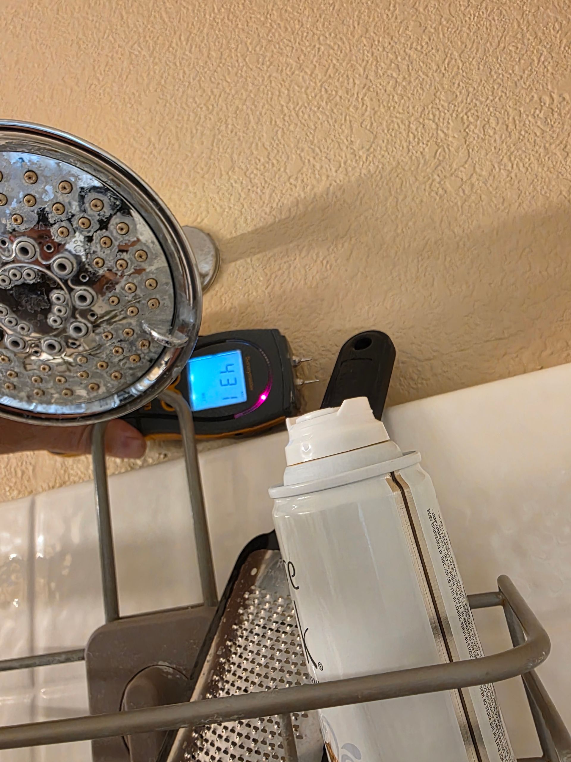 Showerhead held over a water meter in a bathroom. A shower caddy holds a grater and a water filter.