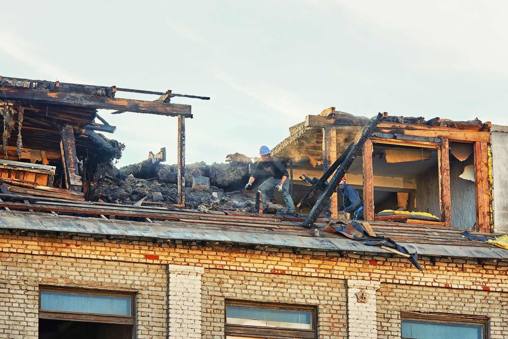 Damaged brick building with burnt roof; people surveying the damage under a clear sky.