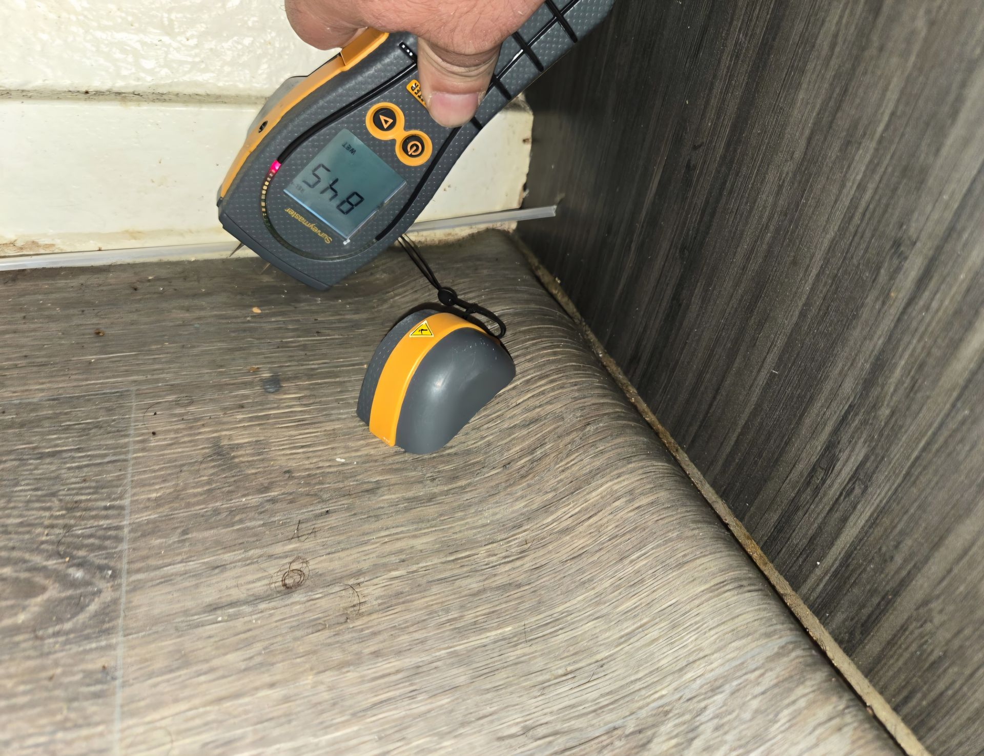 Person using a moisture meter on a damaged floor near a wall.