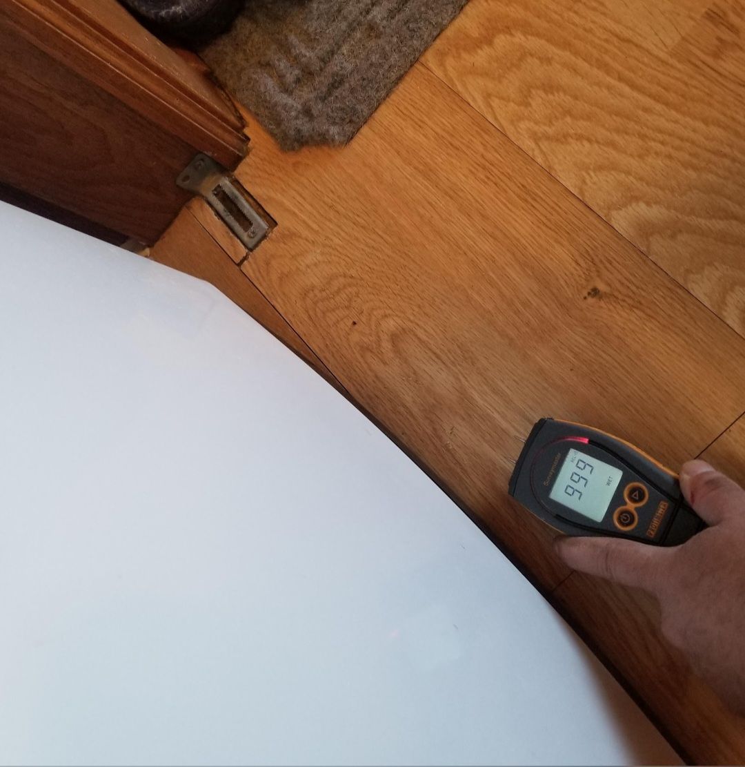 Person holding a moisture meter on a wood floor next to a white appliance.