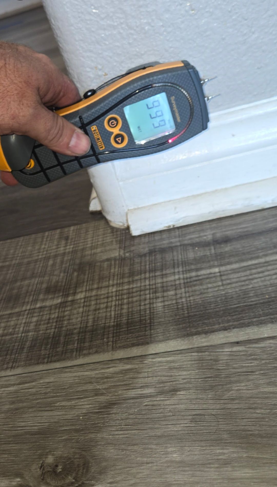 A hand holding a moisture meter on a white baseboard; the meter reads 5.6%, next to wood flooring.