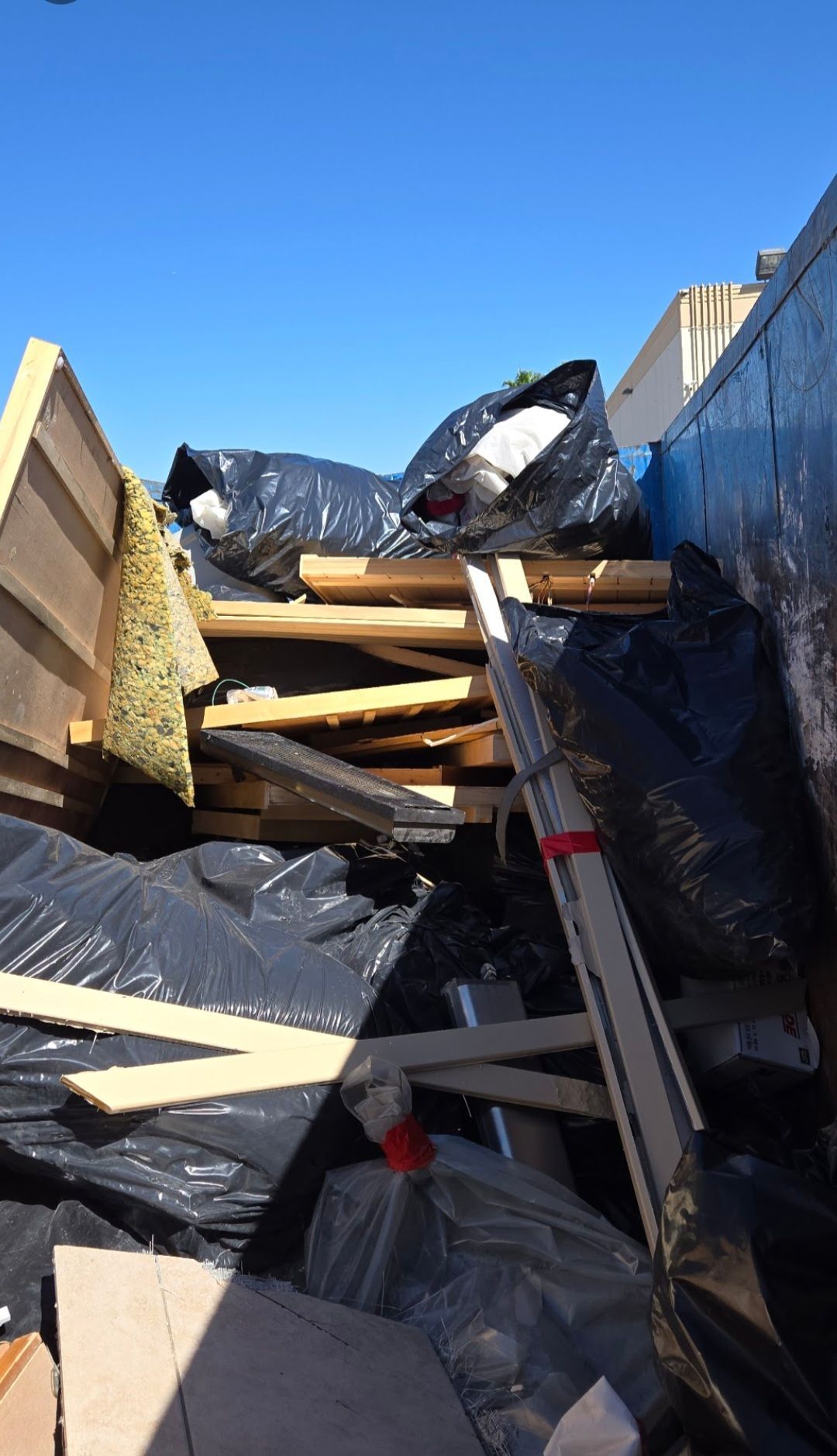 A dumpster filled with debris including black bags, wood, and insulation under a blue sky.