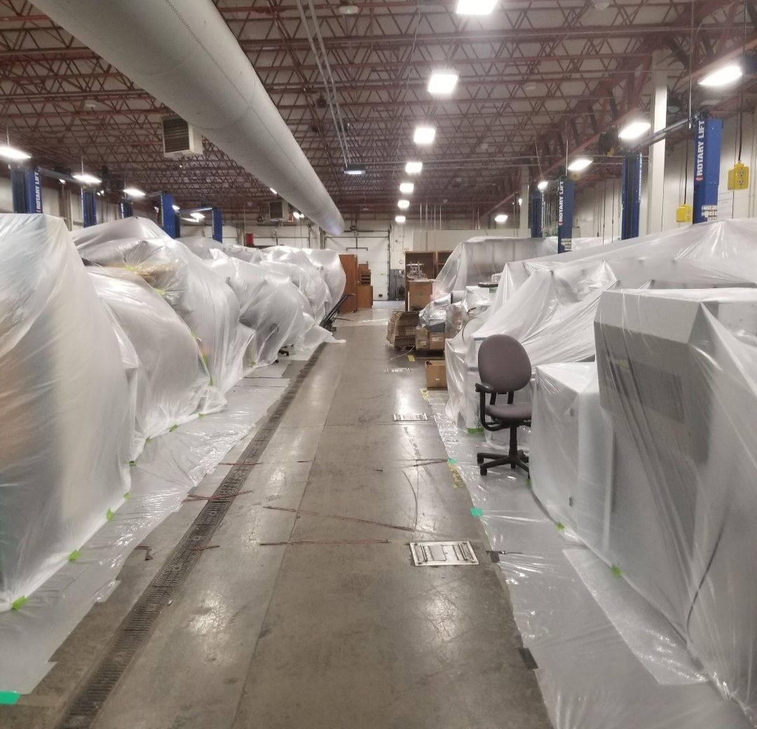 Warehouse interior with equipment covered in plastic, a gray floor, and overhead lighting.