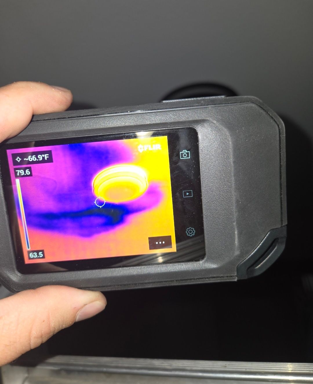 Hand holding a thermal camera, showing a hot, yellow-orange object against a cool, purple-blue background.