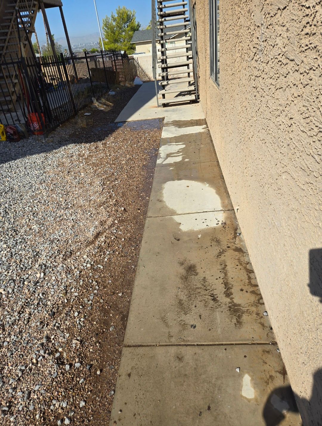 Concrete walkway next to a tan building and gravel yard. A metal staircase is in the background.