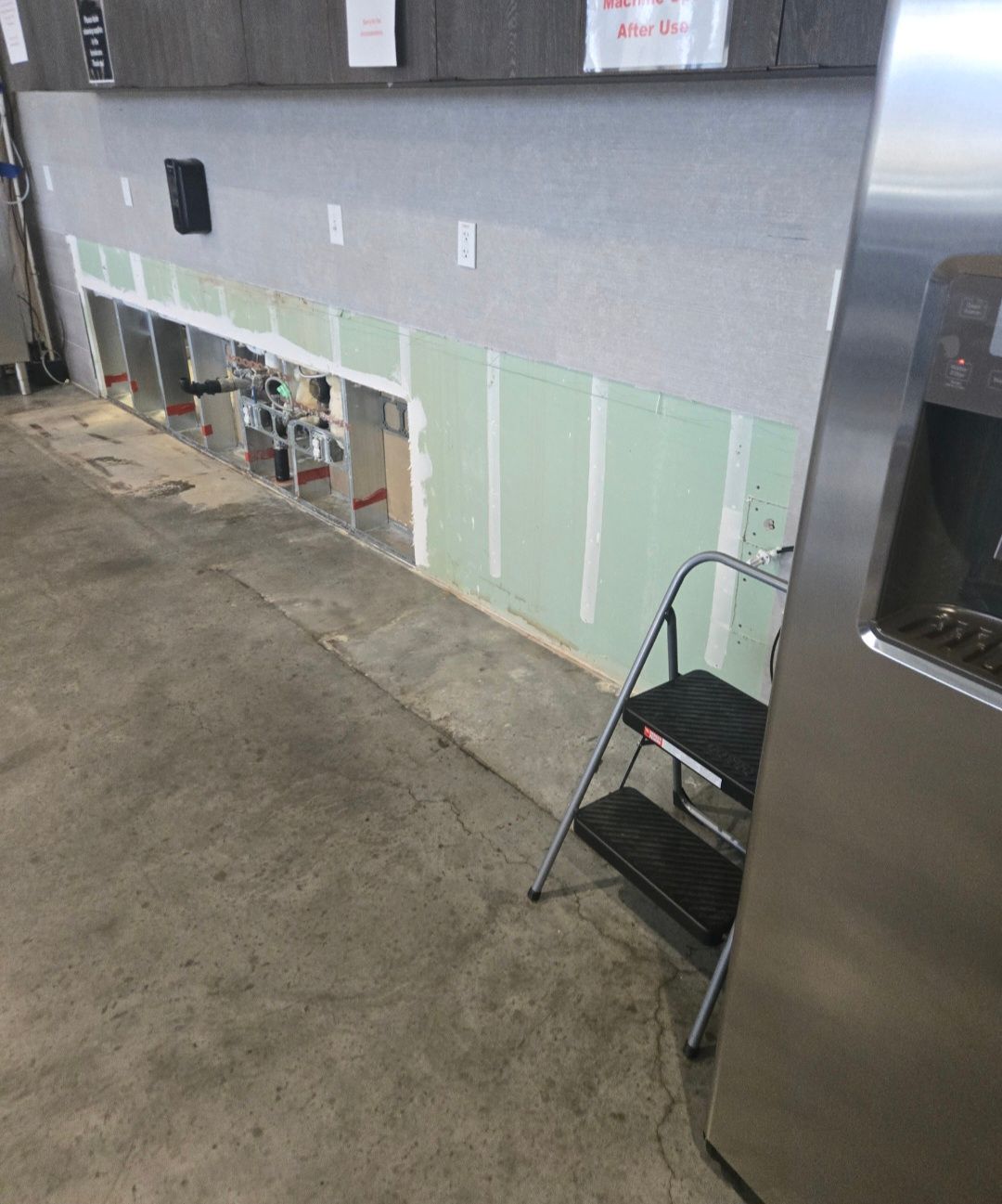 Construction site with exposed wall studs, drywall, and a stainless steel cooler. Stepladder nearby.