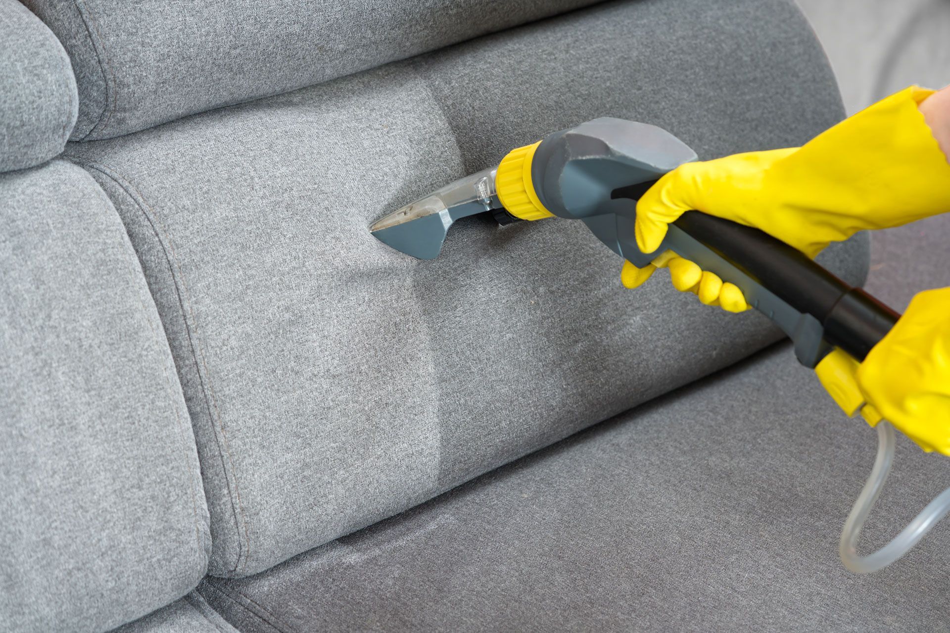 Person in yellow gloves cleaning a gray couch with a handheld upholstery cleaner.