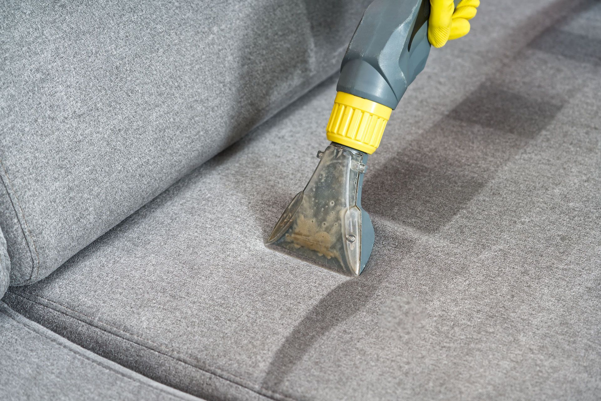 Upholstery being cleaned with a handheld steam cleaner; gray couch, yellow-accented device.
