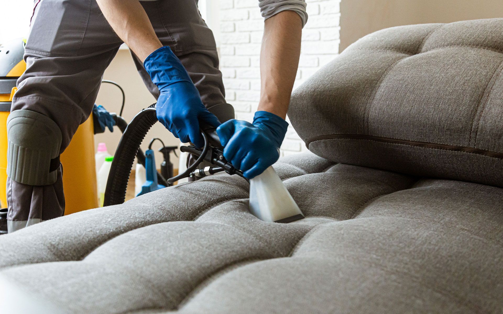 Person in blue gloves cleaning a gray couch with a vacuum cleaner.