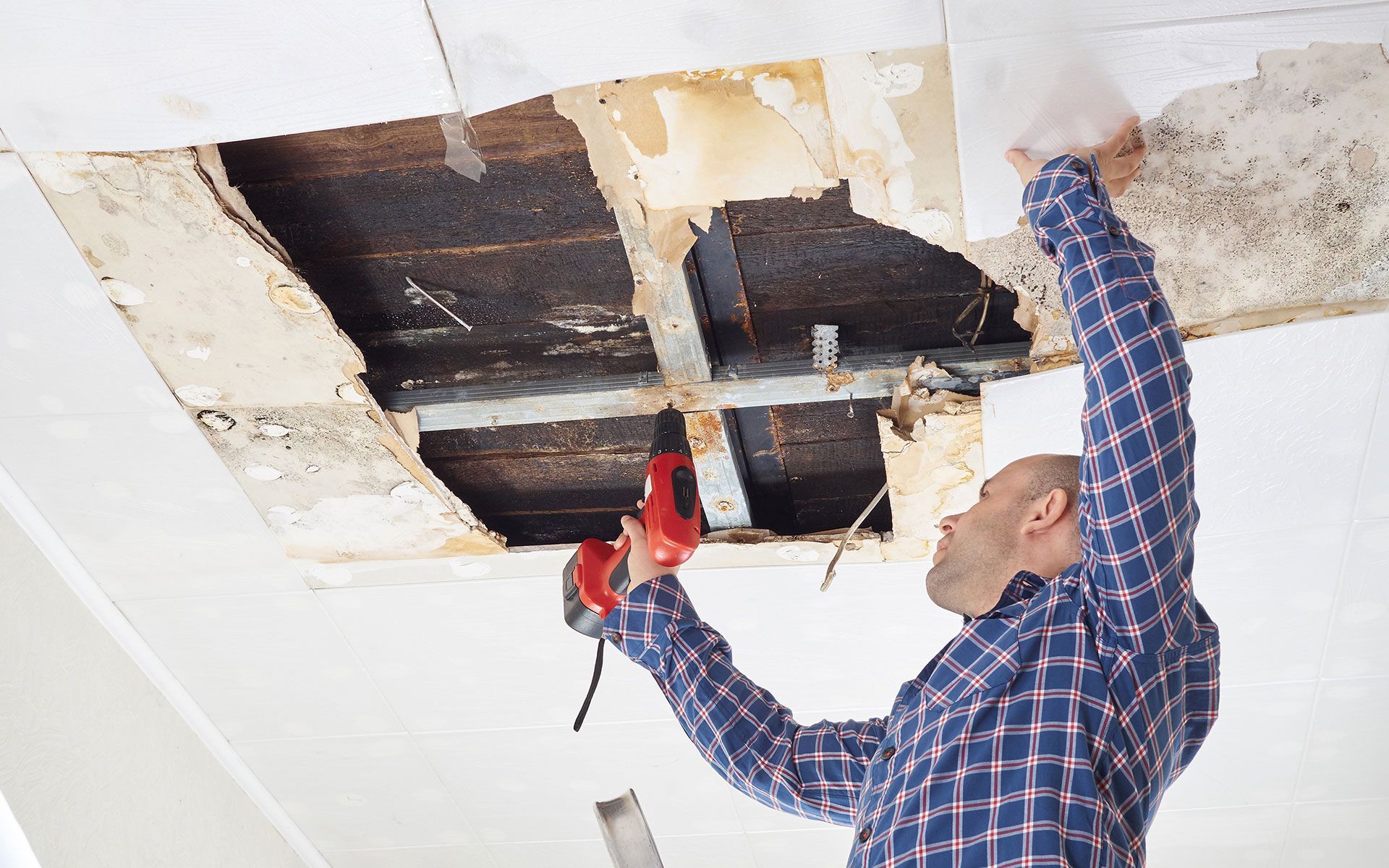 Man repairs damaged ceiling, holding a drill, working in a room with a visible hole.