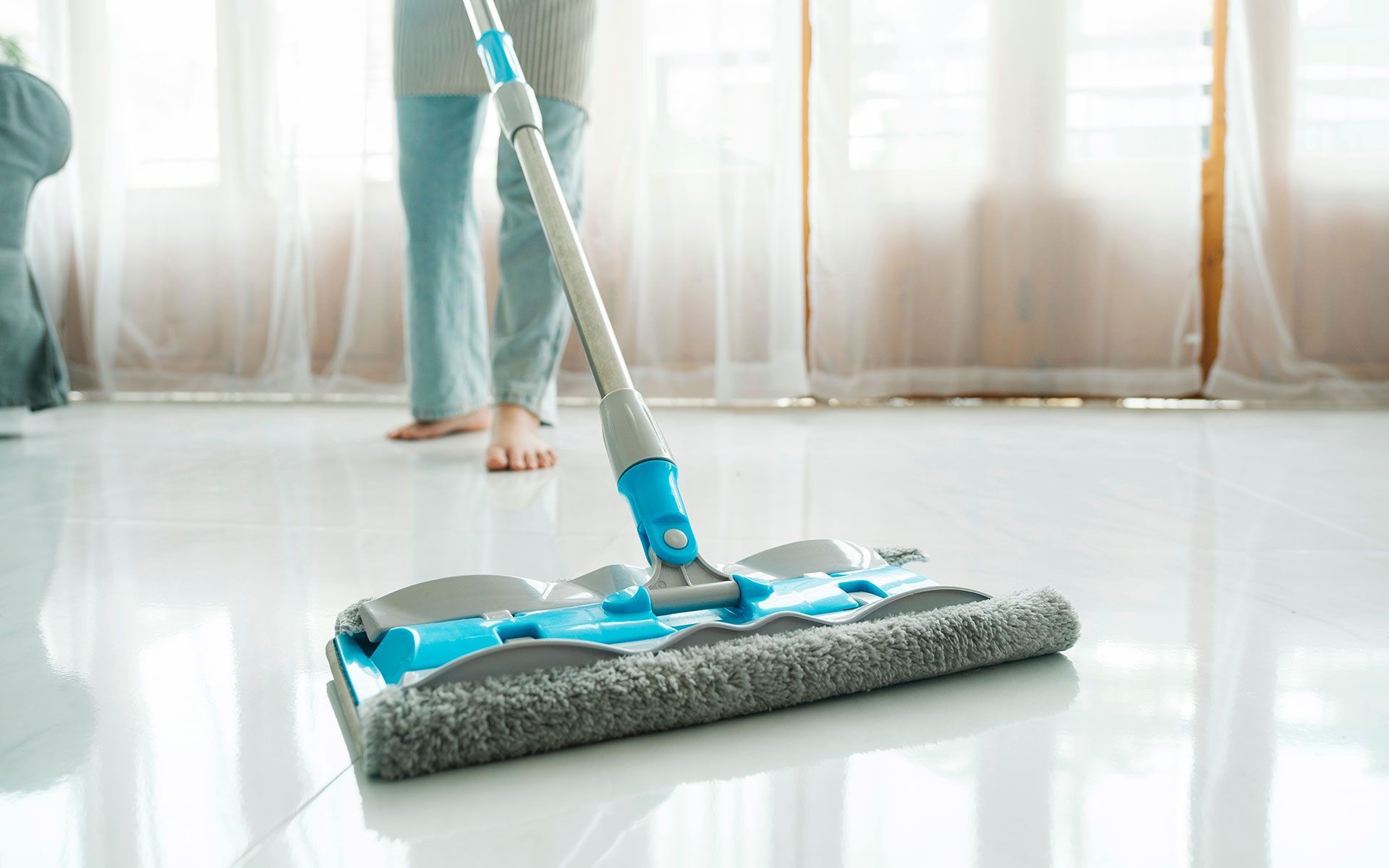 Person mopping a shiny floor with a blue and gray mop in a sunlit room.