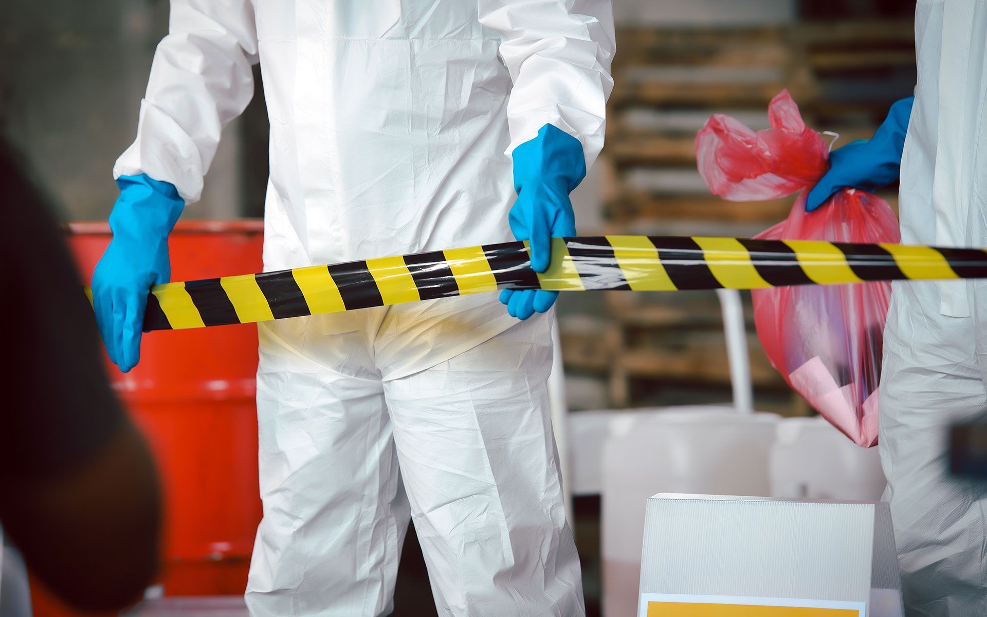 People in hazmat suits holding caution tape in a warehouse; a red barrel is visible.