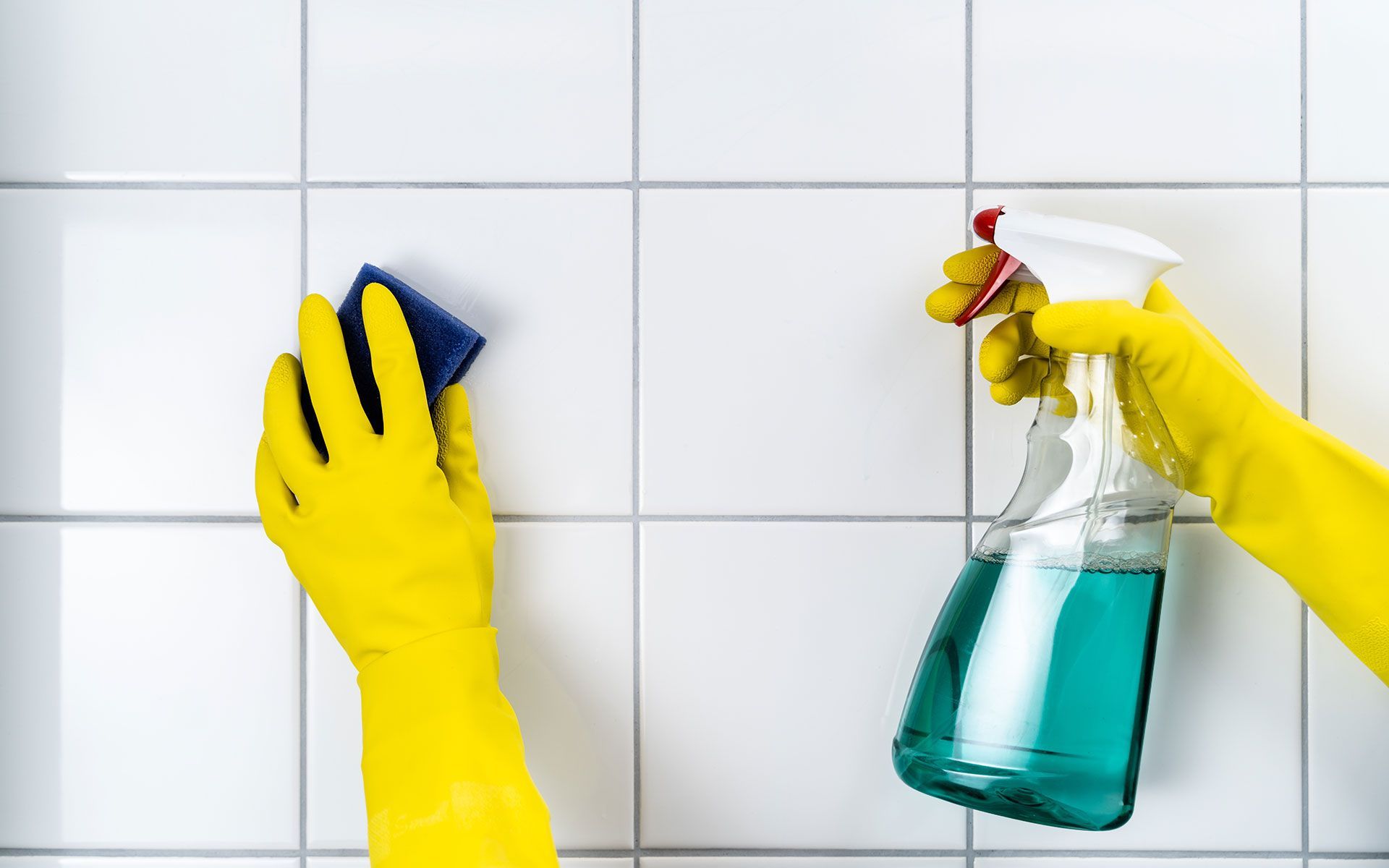 Person wearing yellow gloves cleaning white tiled wall with a spray bottle and sponge.