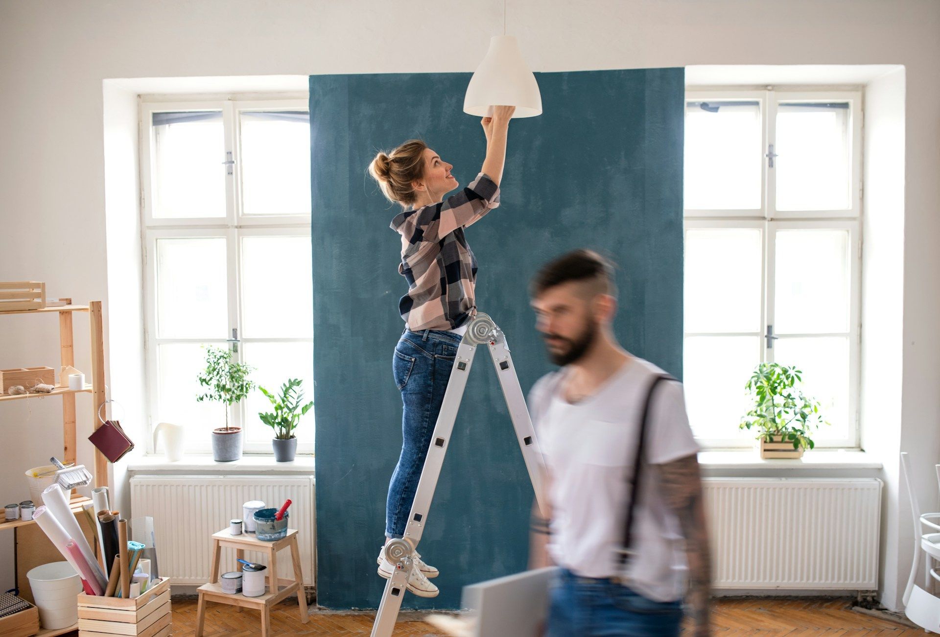 A woman is standing on a ladder hanging a lamp while a man looks on.