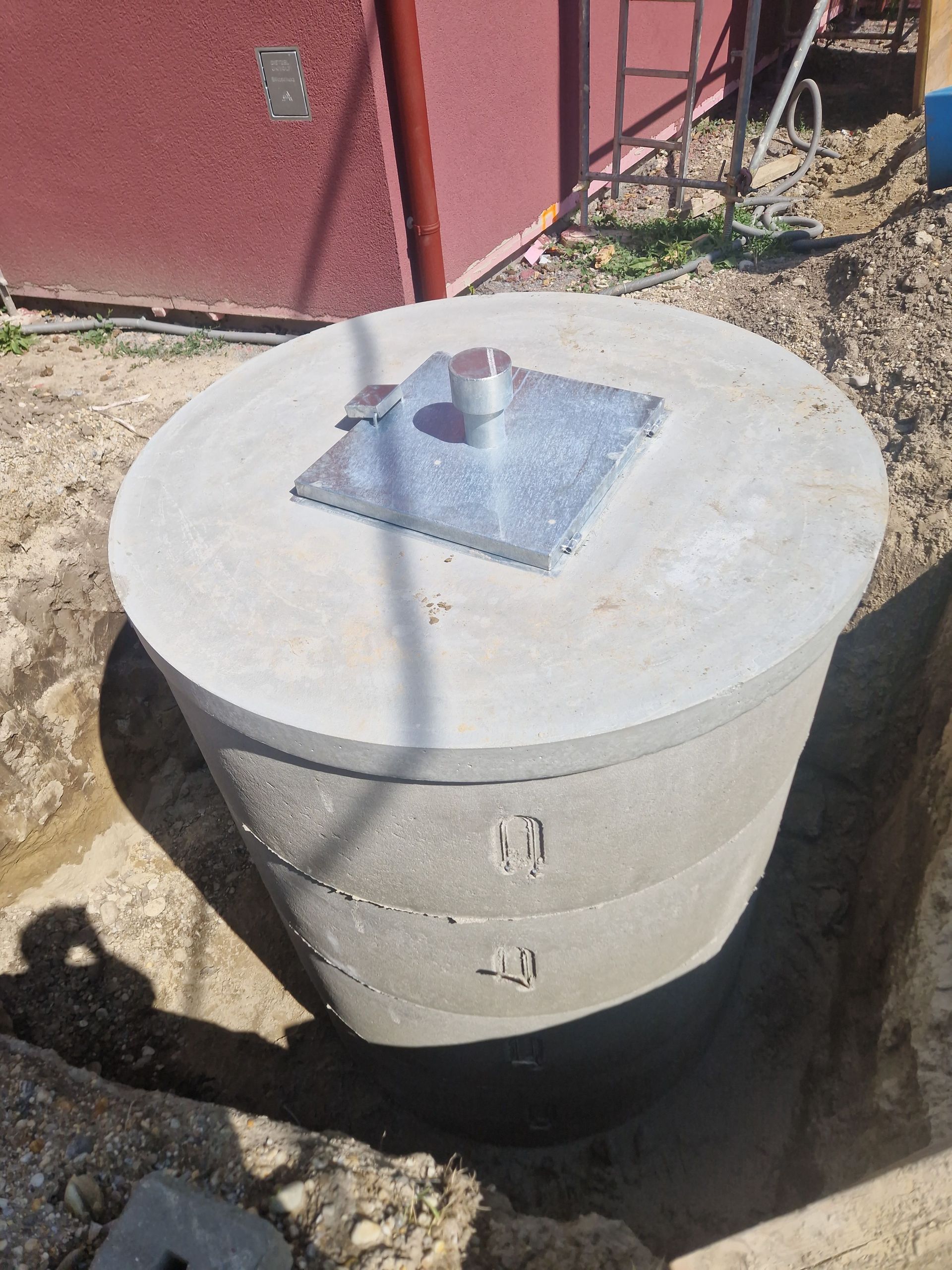 Eine zylindrische Klärgrubenkonstruktion aus Beton in einem Erdgraben mit einer Zugangsklappe aus Metall auf dem Deckel.