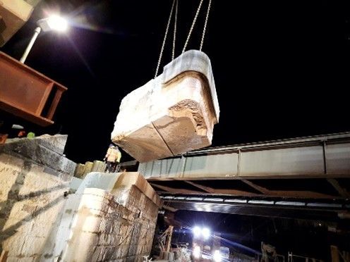 Ein großer Steinblock hängt nachts mit einem Kran über einer Brückenbaustelle.