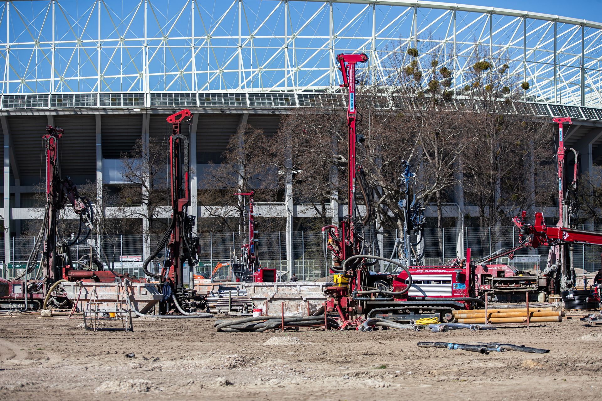 Mehrere rote Bohrgeräte stehen auf einer unbefestigten Baustelle vor einem Stadiongebäude unter strahlend blauem Himmel.