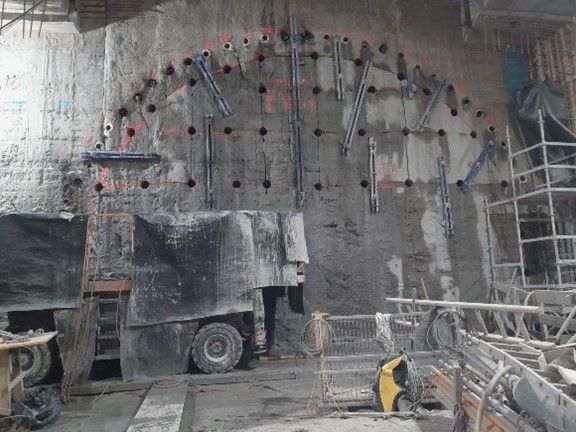 Eine Tunnelortbrust mit Bohrlöchern und Stützankern, auf der ein schweres Bohrgerät im Baustellenumfeld zu sehen ist.