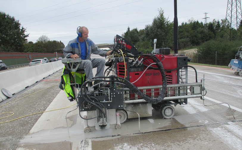 Ein Arbeiter mit Gehörschutz bedient eine rote, schwere Betonschneidmaschine auf einer Autobahnbaustelle.